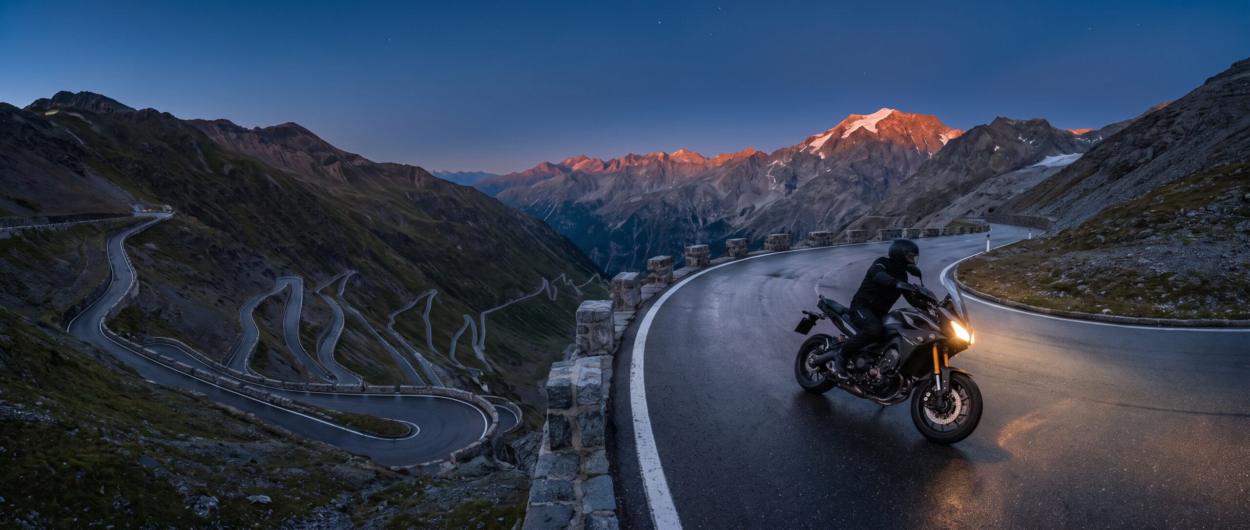A motorcyclist rides a hairpin bend at dusk, headlight on, above a valley of stacked switchbacks