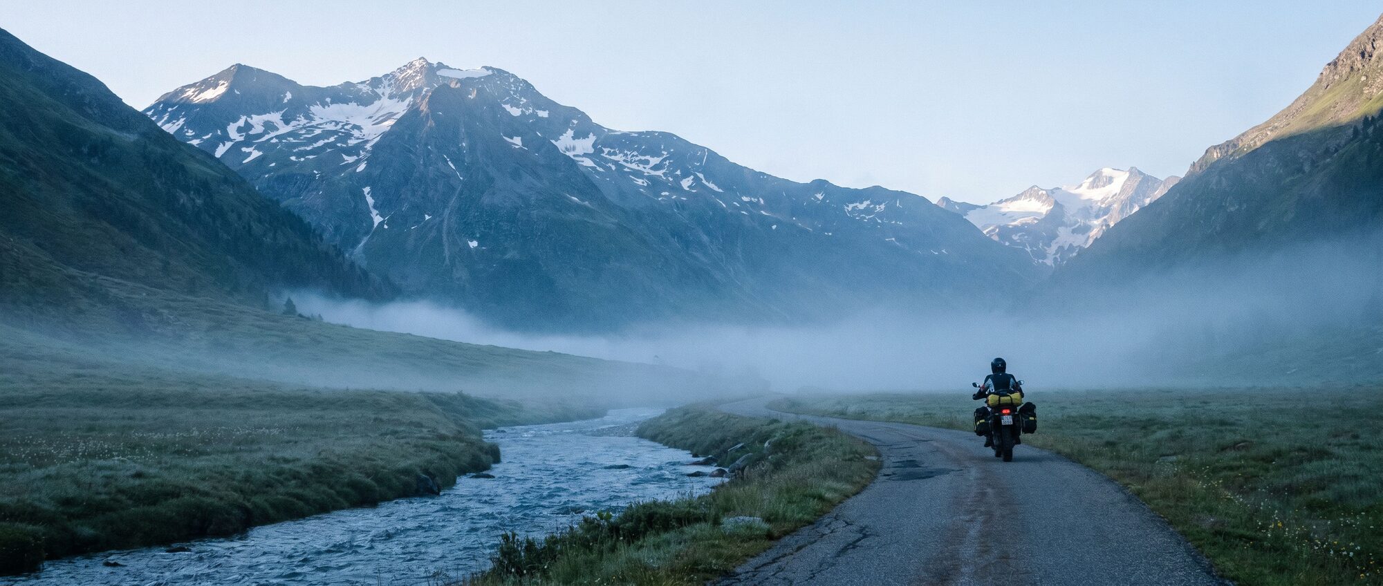 A lone motorcyclist rides a narrow road through a remote foggy alpine valley at dawn
